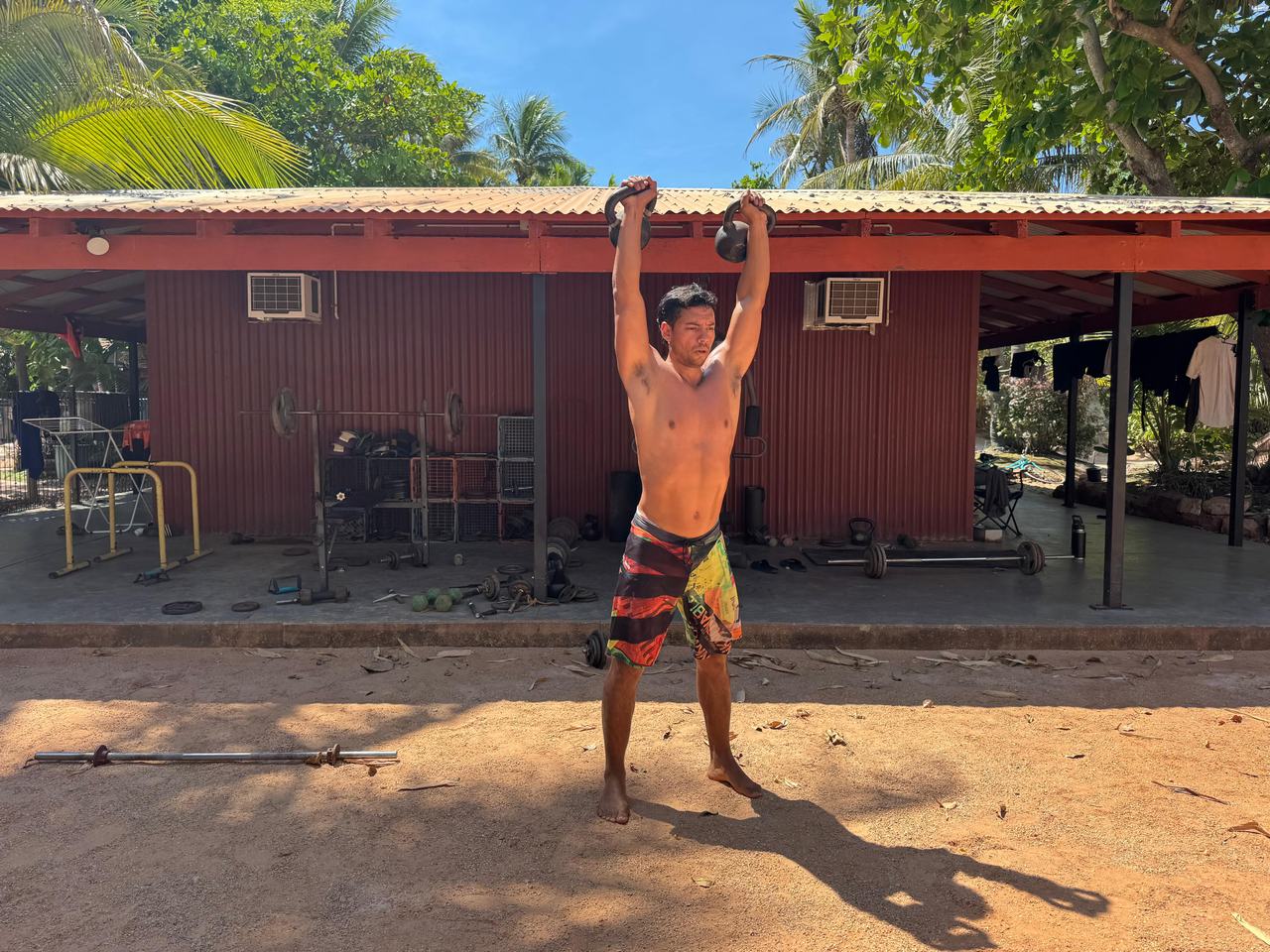 Bruno distende due kettlebell sopra la testa in un cortile, davanti a una baracca in lamiera con i pesi sparsi sotto la tettoia